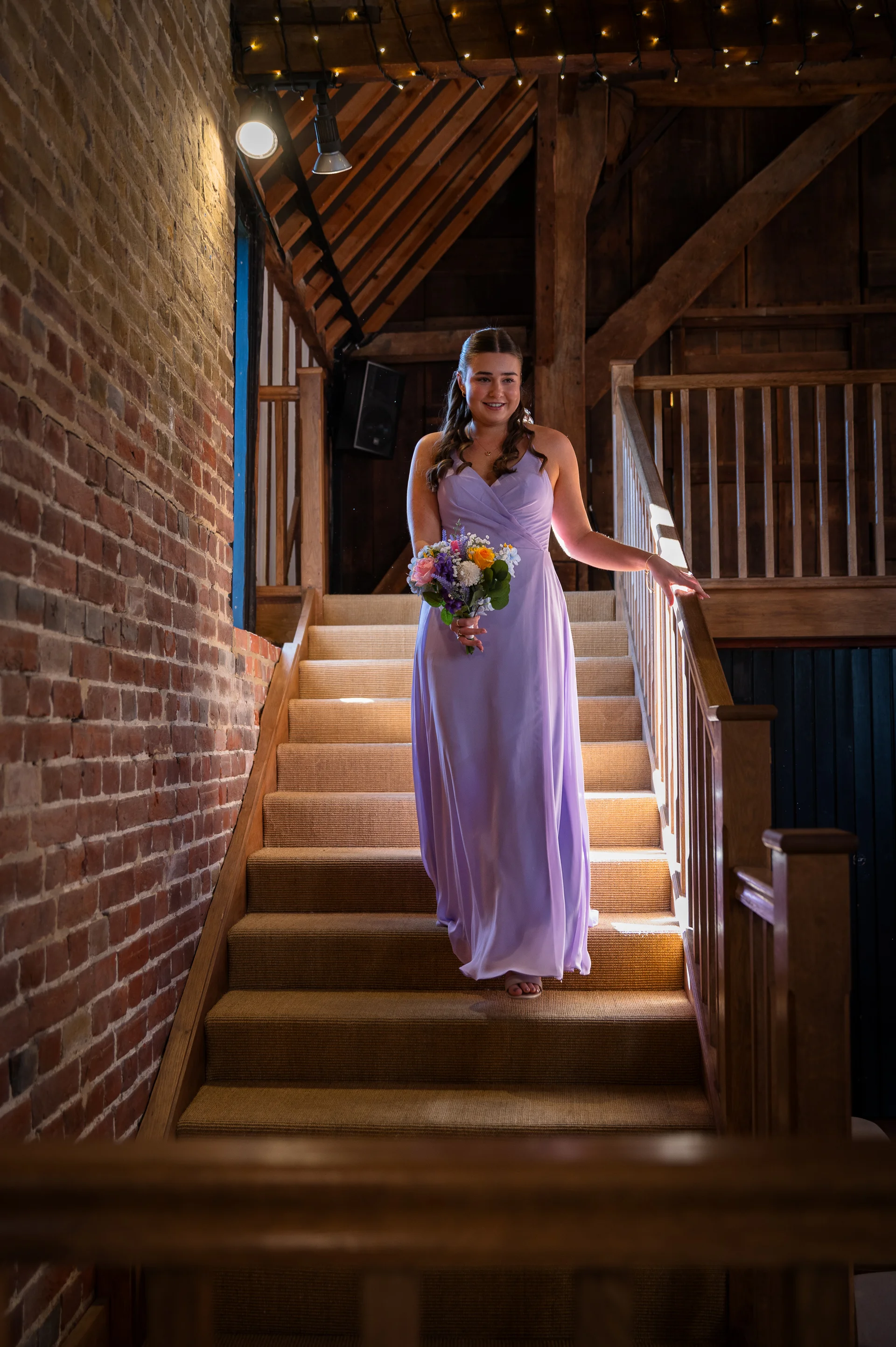 A bridesmaid in a lilac dress descends a wooden staircase inside an oak-framed rustic barn, holding a colourful bouquet.