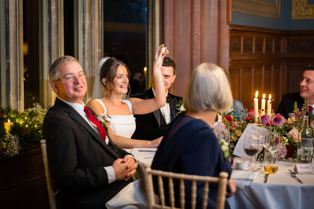 A joyful bride raises a pocket watch in the air during the wedding breakfast while seated with guests at a candlelit top table.