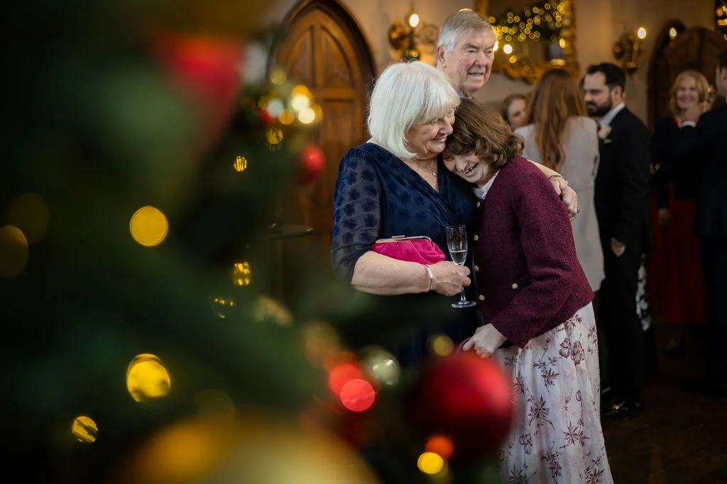 An older woman and another wedding guest share a tender, emotional embrace near a softly blurred Christmas tree during the wedding drinks reception.