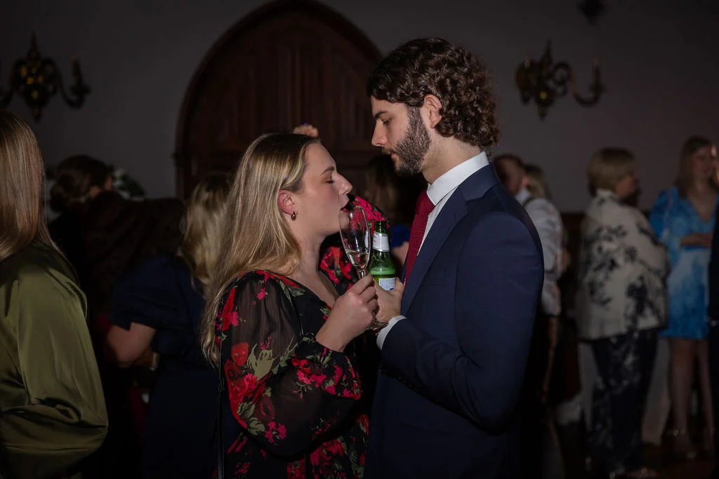 A man in a navy suit and red tie shares a close, tender moment on the dance floor with a guest in a floral print dress holding a glass of champagne.