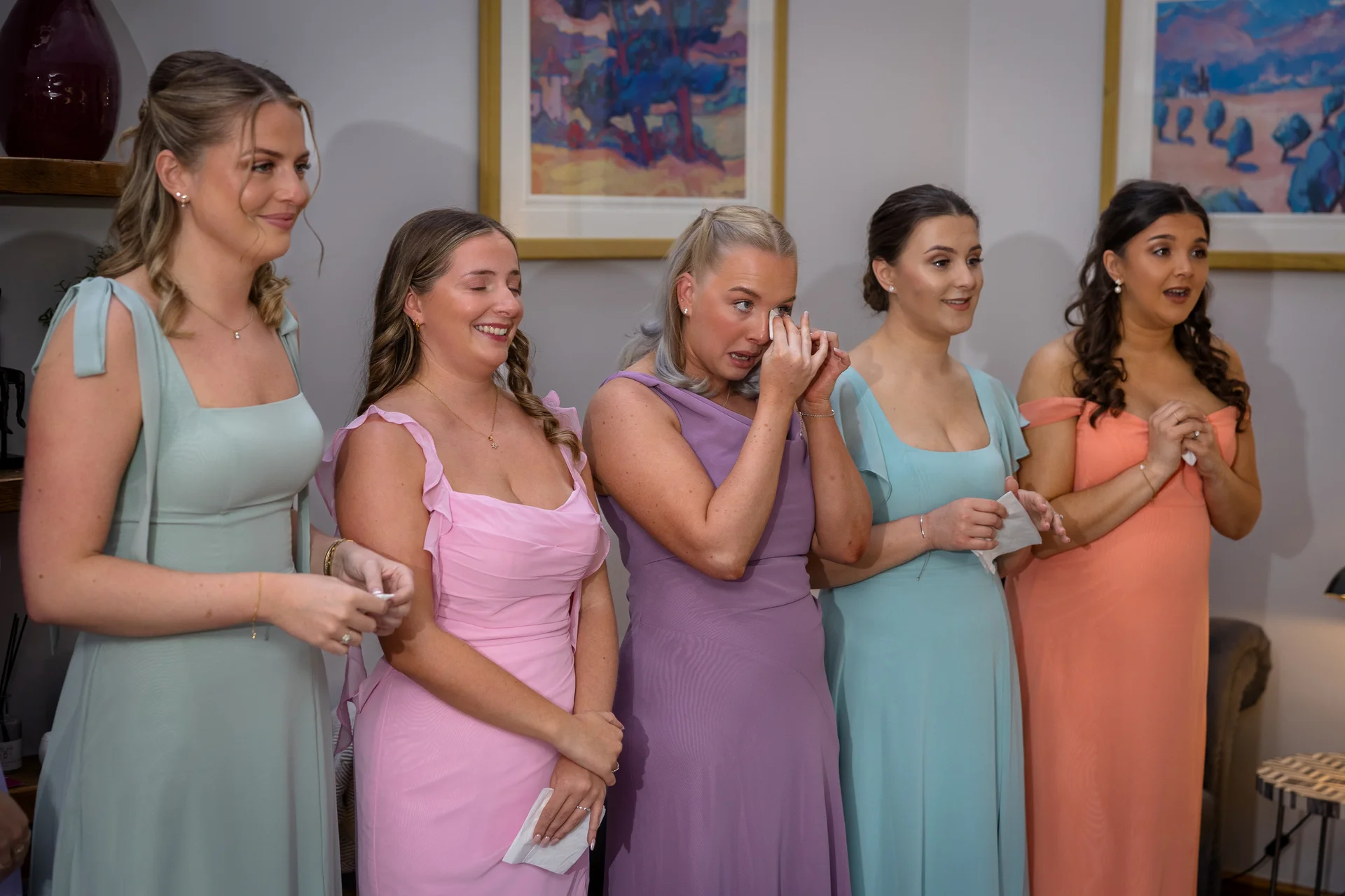 A group of bridesmaids in colourful pastel dresses wipe away happy tears while watching wedding speeches inside an art gallery venue.