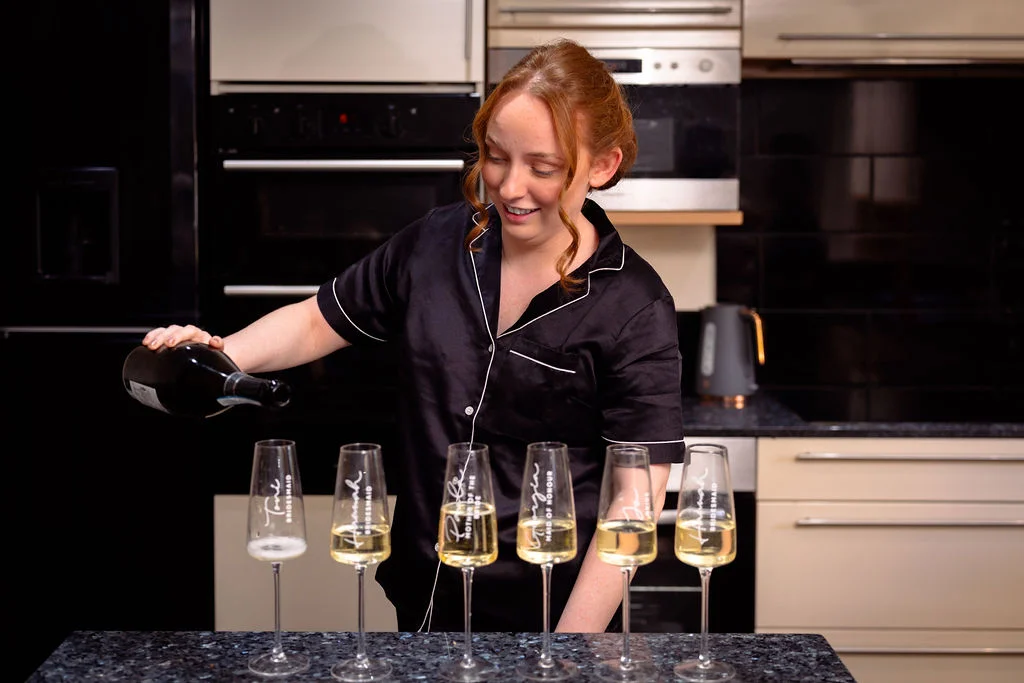 A smiling woman with red hair wearing matching black pyjamas pours champagne into a line of personalised flutes during morning wedding preparations.
