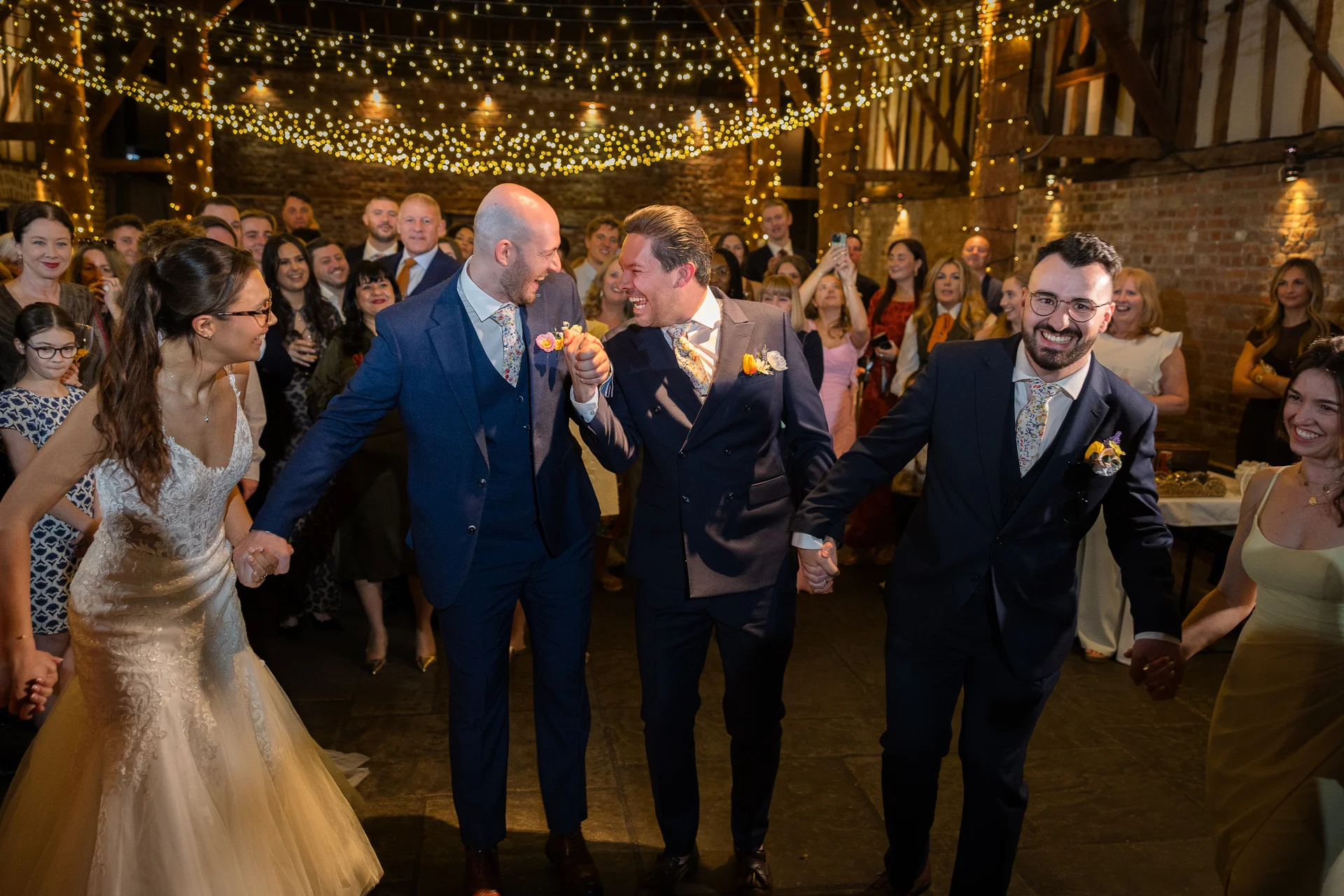A joyful couple and their wedding party holding hands and laughing together in a rustic barn illuminated by a glowing canopy of fairy lights.
