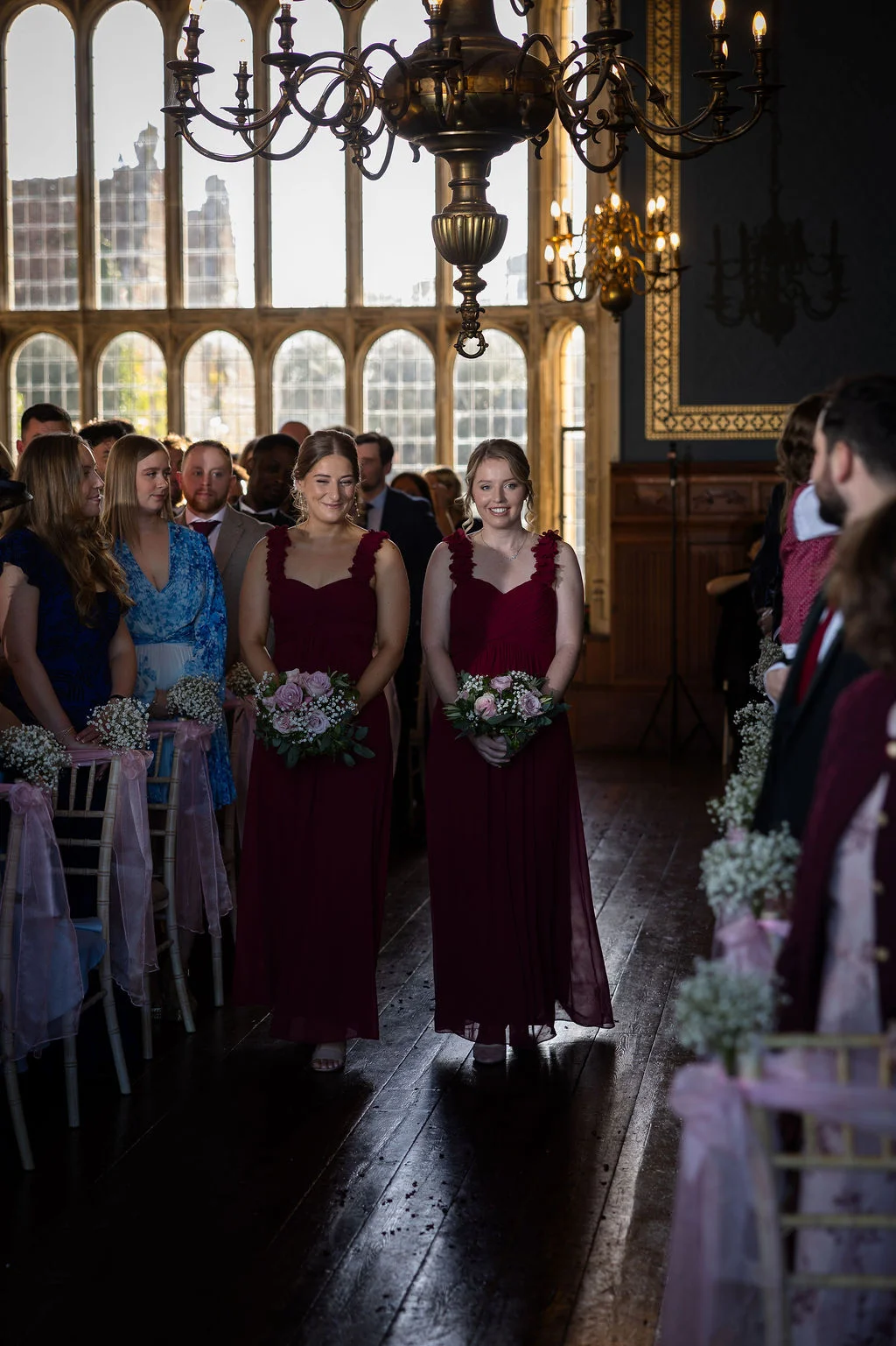 Two smiling bridesmaids in matching burgundy dresses walk down the aisle holding bouquets during a ceremony inside a grand hall with large windows.