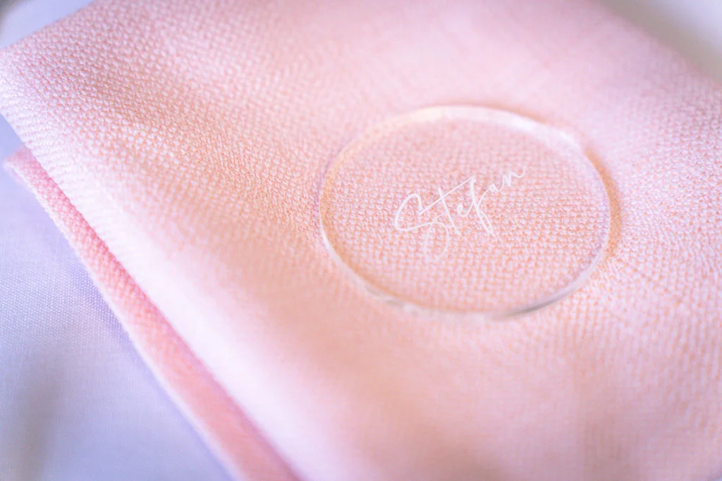 A close-up detail shot shows a round clear acrylic place card featuring modern calligraphy resting on top of a folded pink fabric napkin.