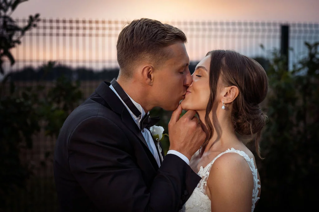 A bride and groom kissing in front of a fence at sunset.