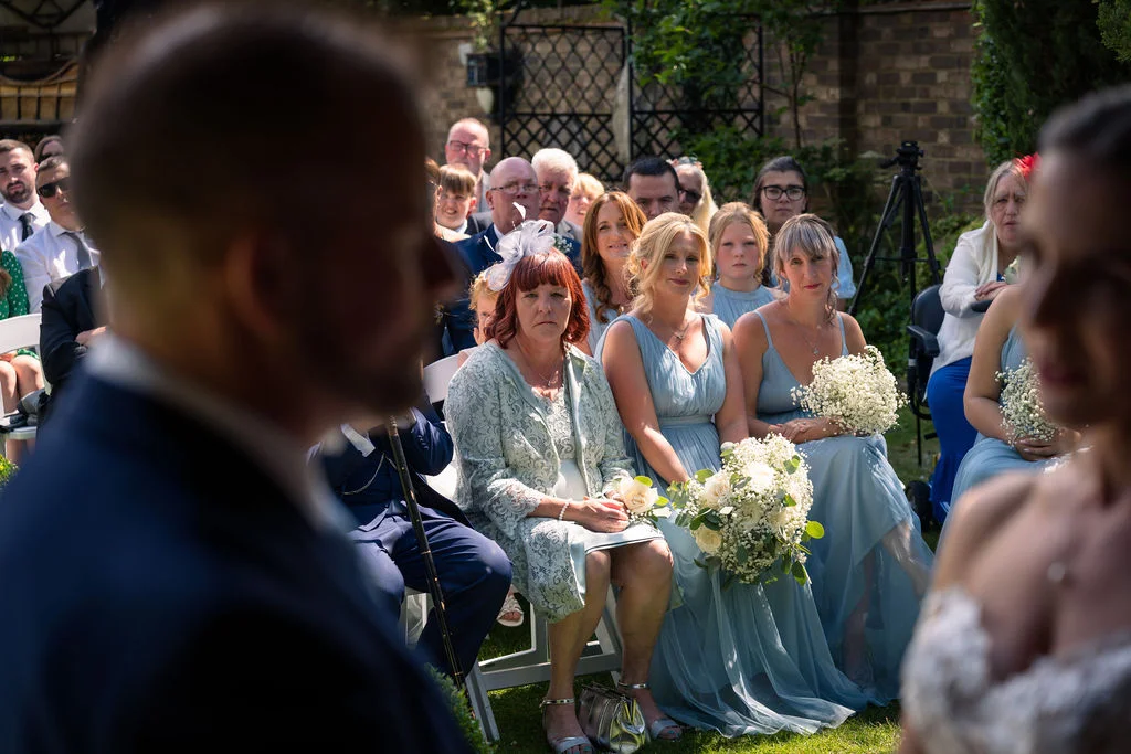 An over-the-shoulder shot captures the groom standing at the outdoor ceremony as guests and bridesmaids in dusty blue dresses look on smiling.