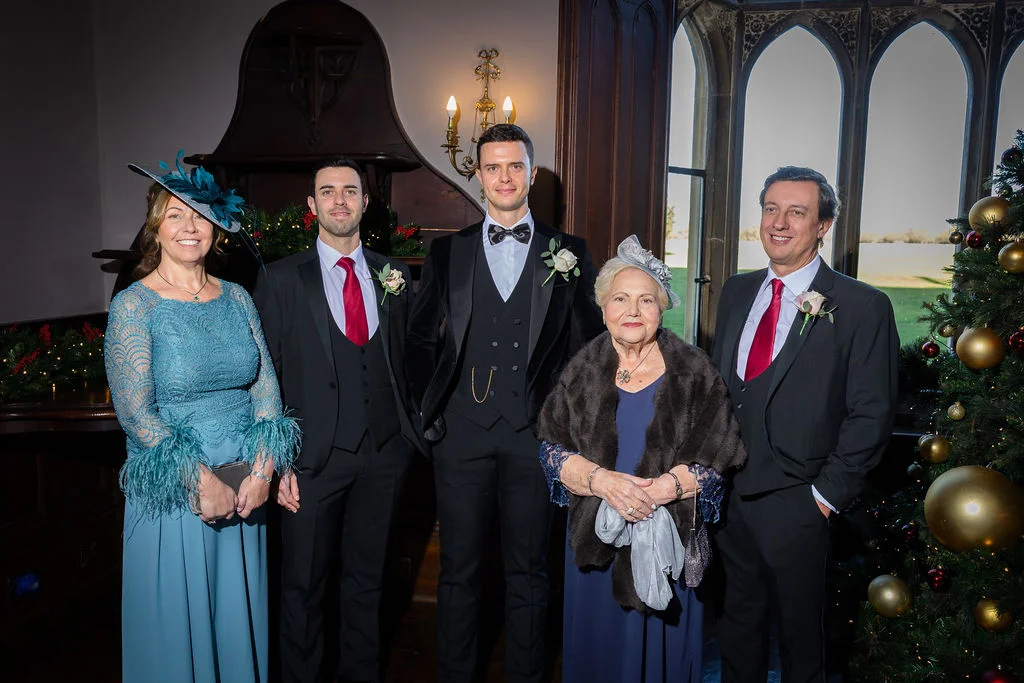 A group of family members pose for a formal portrait inside a stately home during a festive Christmas wedding, featuring a Christmas tree and fireplace.