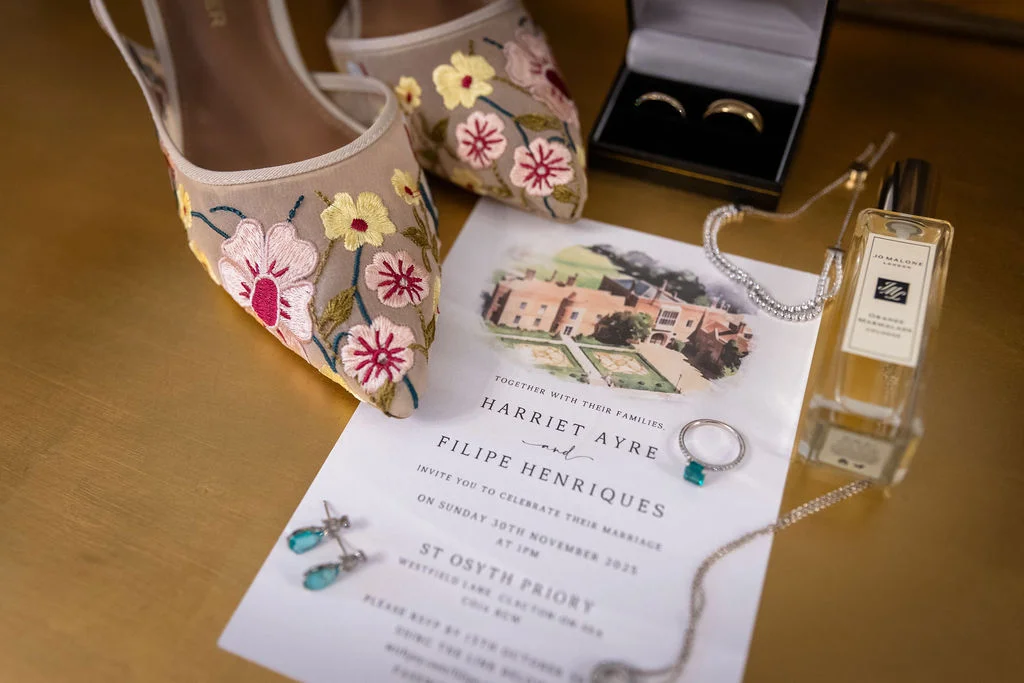 An elegant flat lay captures a bridal invitation suite featuring a venue illustration, accompanied by embroidered floral low heels, delicate drop earrings, wedding bands in a box, and a bottle of perfume.