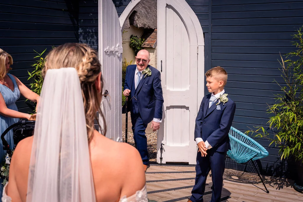 An over the shoulder shot captures a bride in a veil and braided bun looking toward her smiling father and young page boy stepping through a white arched doorway.