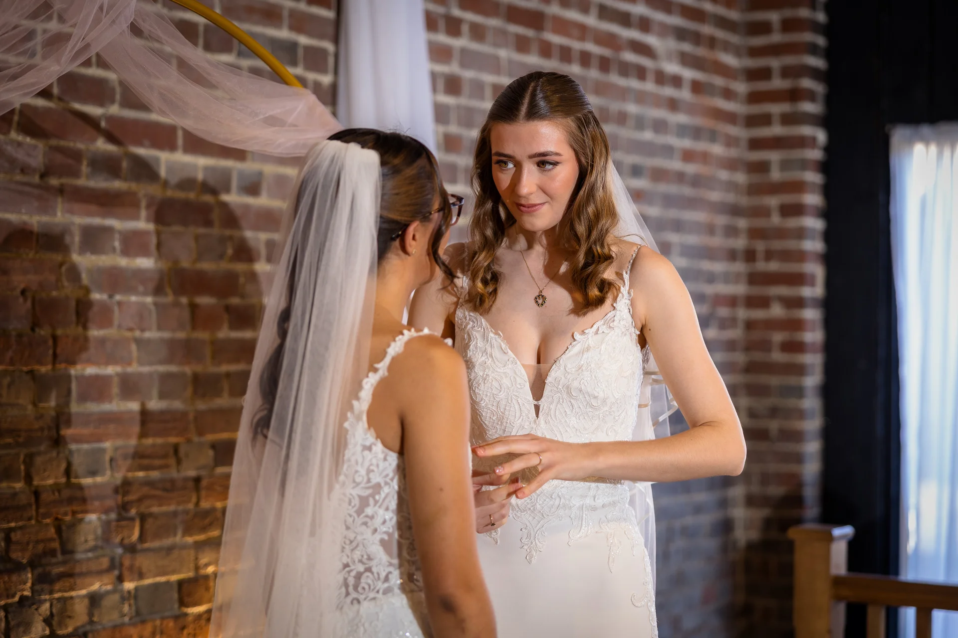 Two brides in lace wedding dresses lovingly exchange rings during their indoor ceremony against an exposed brick wall.