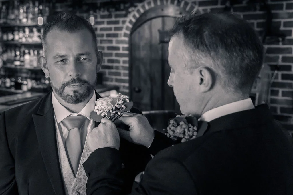 A black and white photograph of one groom attentively pinning a floral buttonhole onto the lapel of another groom inside a rustic pub.