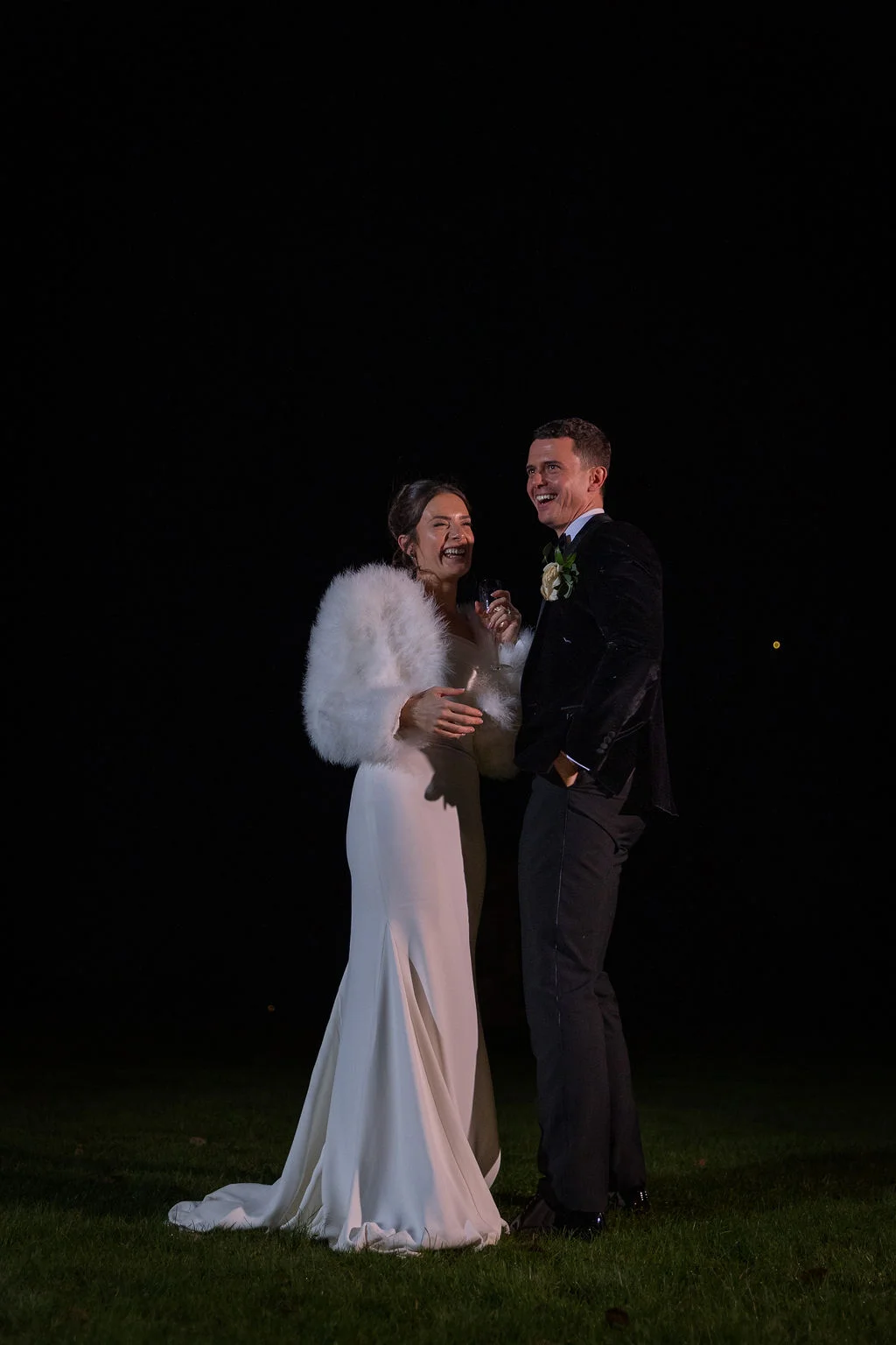 Newlyweds laughing together outdoors at night, the bride wearing a sleek gown with a thigh split and a fluffy white stole while holding a drink.