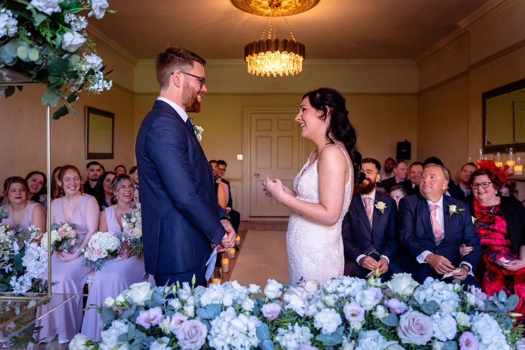 The bride and groom exchange vows and rings during a bright indoor civil ceremony surrounded by seated guests and lush floral arrangements.
