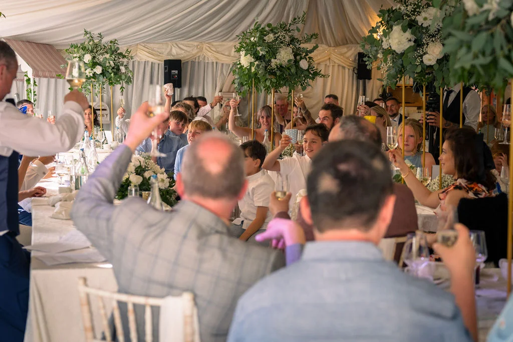 Wedding guests raise their glasses for a toast during the wedding breakfast inside a marquee decorated with white draping and tall floral centrepieces.