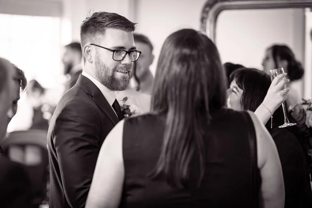 A smiling groom wearing glasses and a suit mingles with guests at a wedding drinks reception while someone raises a glass.