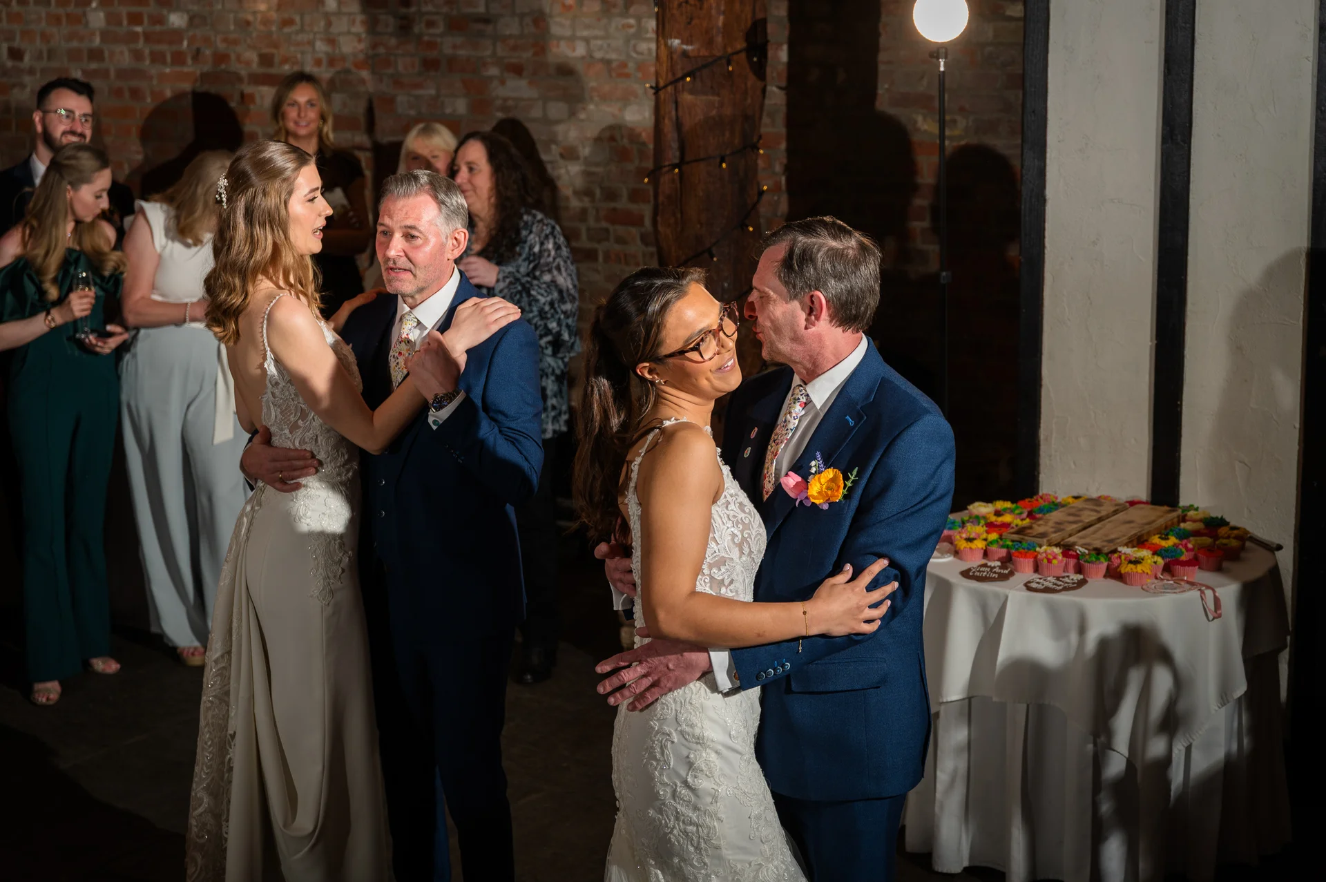 Two couples share a joyous moment dancing together during the evening reception inside a rustic barn venue with exposed brick walls.