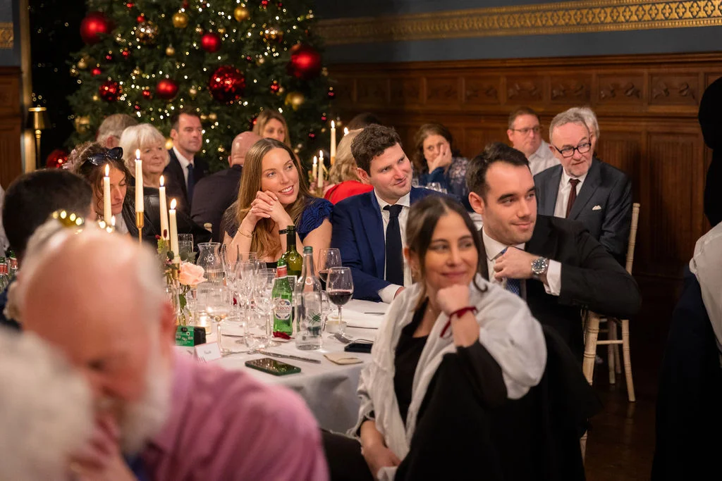 Wedding guests sit together around a candlelit dining table during speeches, with a decorated Christmas tree glowing in the background.