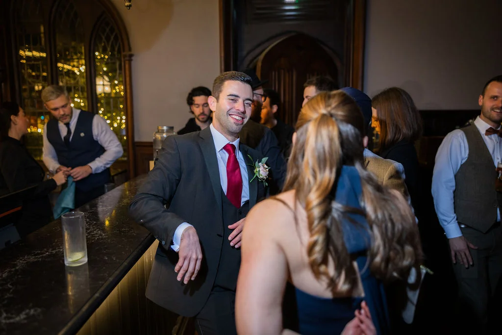 A smiling groom wearing a charcoal three-piece suit with a red tie leans against a pub bar during a lively evening drinks reception.
