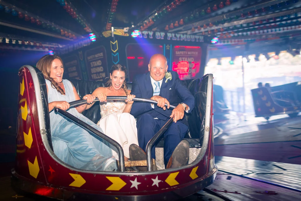 A joyful bride, groom, and guest laugh loudly while riding in a vintage dodgem car at a funfair evening reception.