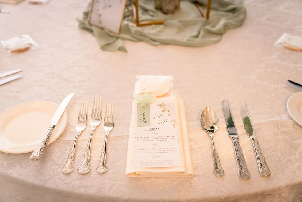 An elegantly set wedding reception table features a printed menu on a folded napkin, silverware, and a sage green table runner.