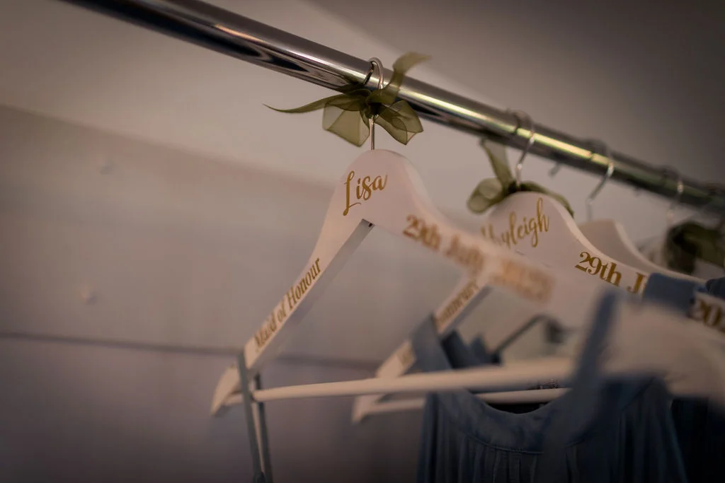 Personalised white wooden hangers featuring gold calligraphy and sage green ribbon bows hang in a row, holding blue bridesmaid dresses.