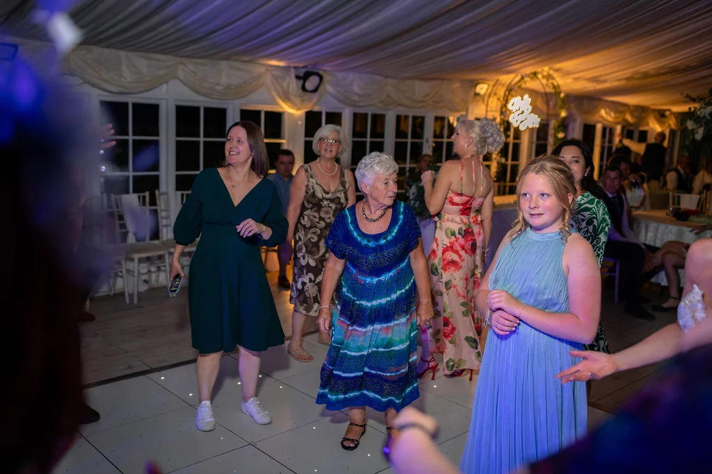 Guests of multiple generations enjoy dancing together on an illuminated dance floor beneath draped marquee ceilings.