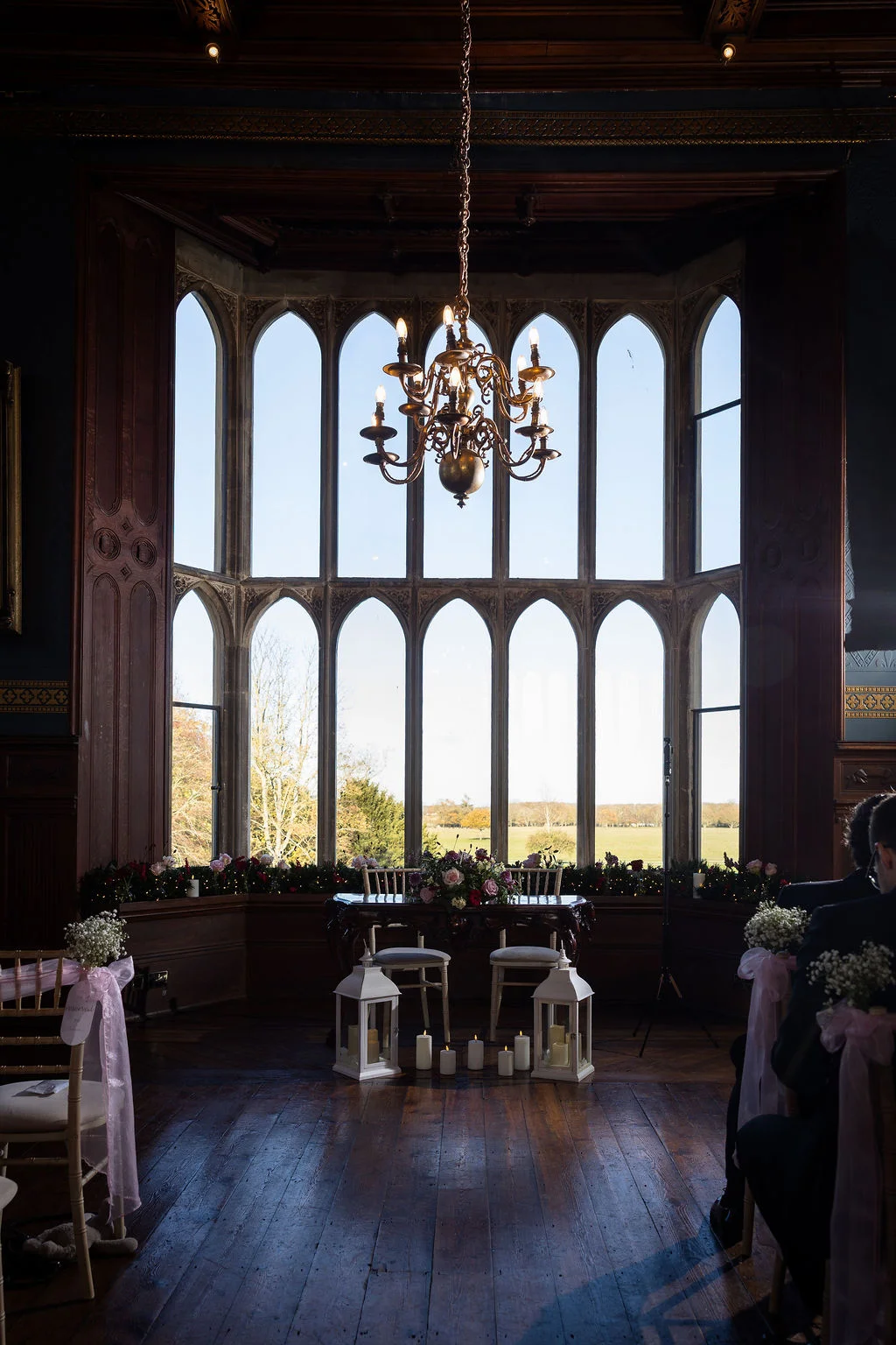 An atmospheric indoor wedding ceremony space featuring a grand arched bay window overlooking a scenic countryside landscape, lit by natural daylight and a central chandelier above the floral-adorned signing table.
