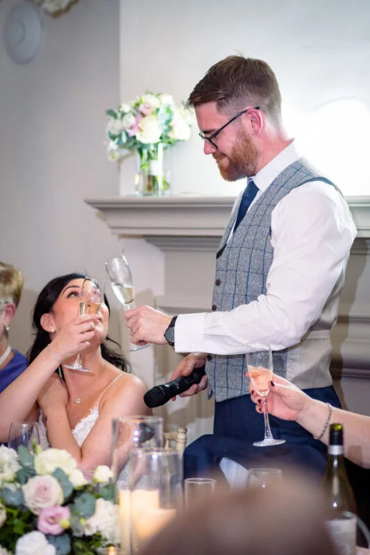 A bearded groom in a tweed waistcoat delivers a speech while clinking champagne glasses with the smiling bride during the wedding breakfast.