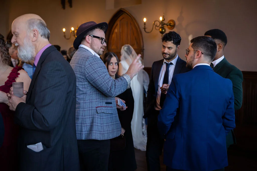A close-up magician in a checked blazer performs a card trick for wedding guests during the drinks reception in a historic hall.