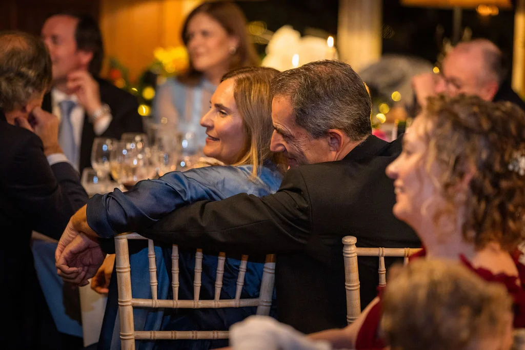 An older couple shares a tender embrace seated at a wedding breakfast table while other guests look on and laugh during the speeches.