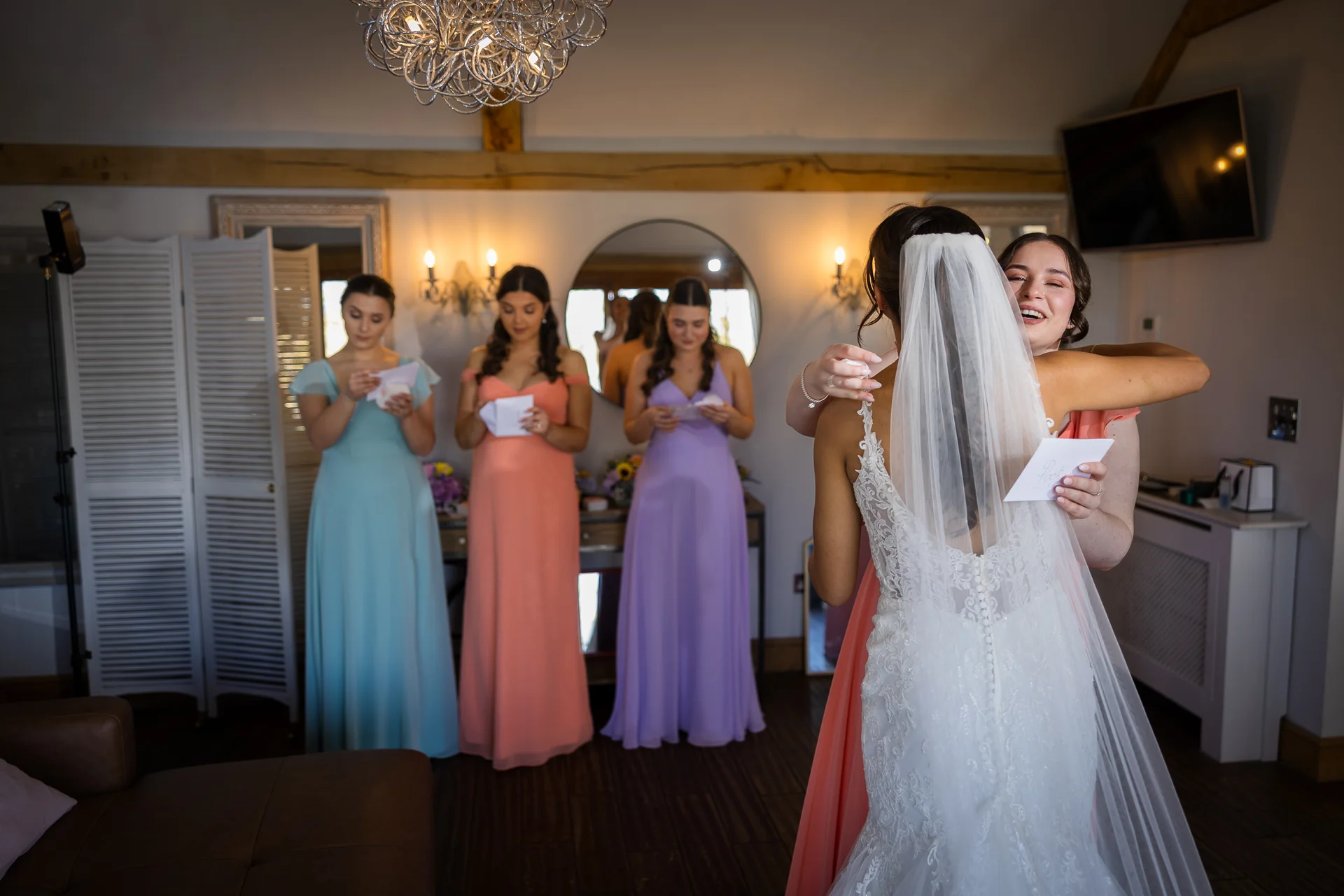 A bride in a lace gown and veil embraces her laughing bridesmaid who is holding a letter, while other bridesmaids in pastel dresses look on during bridal preparations.