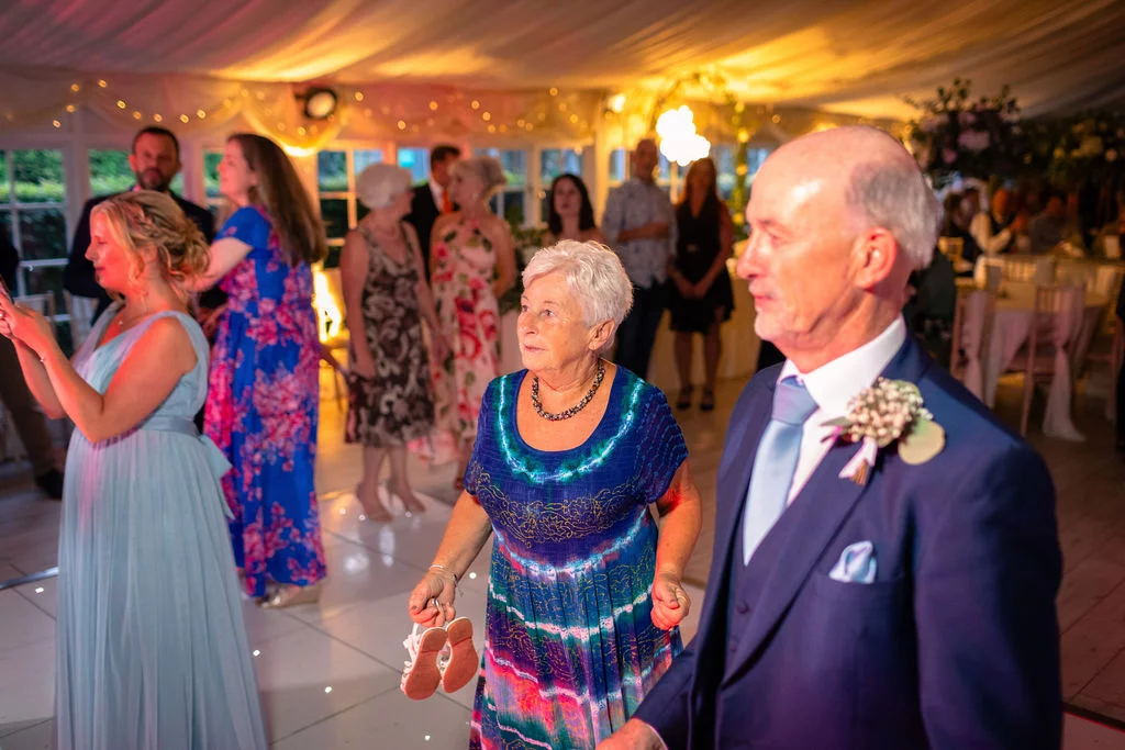 An older man in a navy suit and an older woman in a patterned dress enjoy the evening reception inside a marquee illuminated by warm lighting.