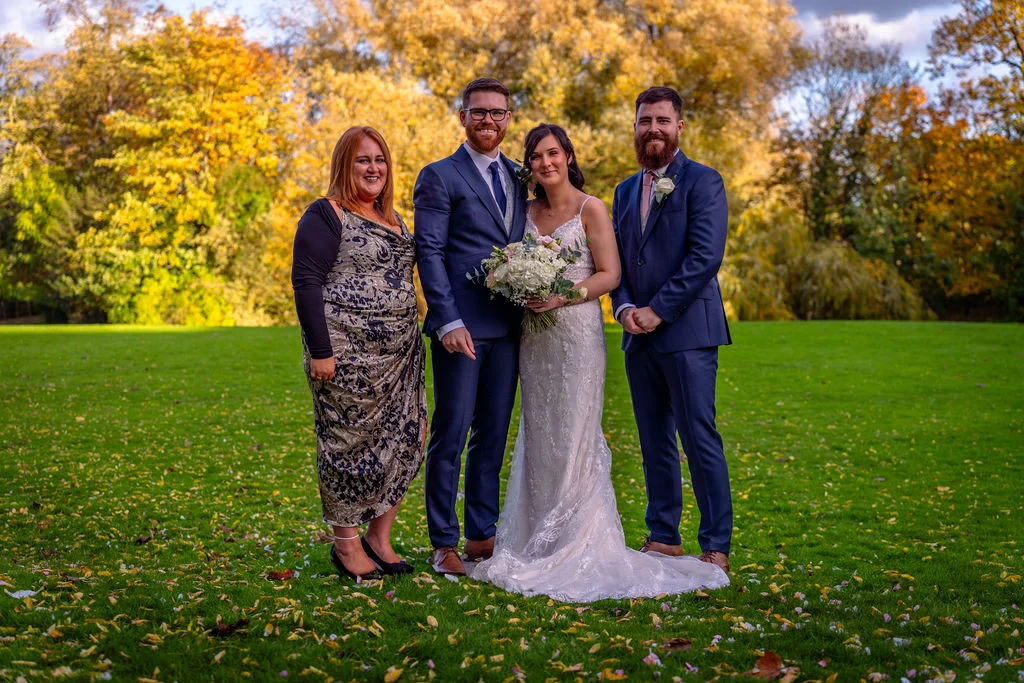 A bridal party poses outdoors on a vibrant green lawn scattered with autumn leaves, surrounded by trees with stunning golden foliage.