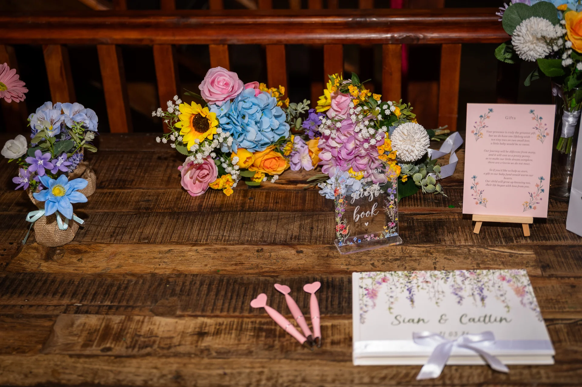A rustic wooden table displays a colourful guest book station featuring vibrant floral arrangements, a small sign, and heart-topped pens.