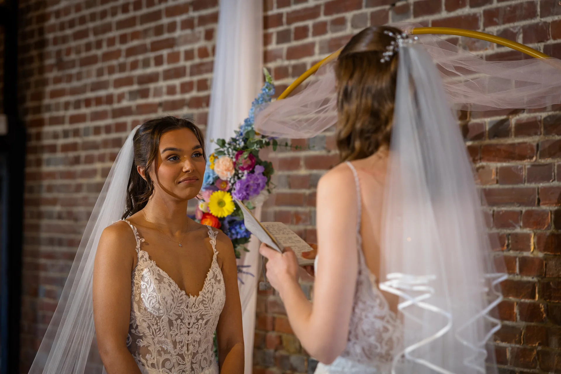 Two brides exchange personal vows during an intimate indoor ceremony framed by an exposed brick wall and a colourful floral archway.