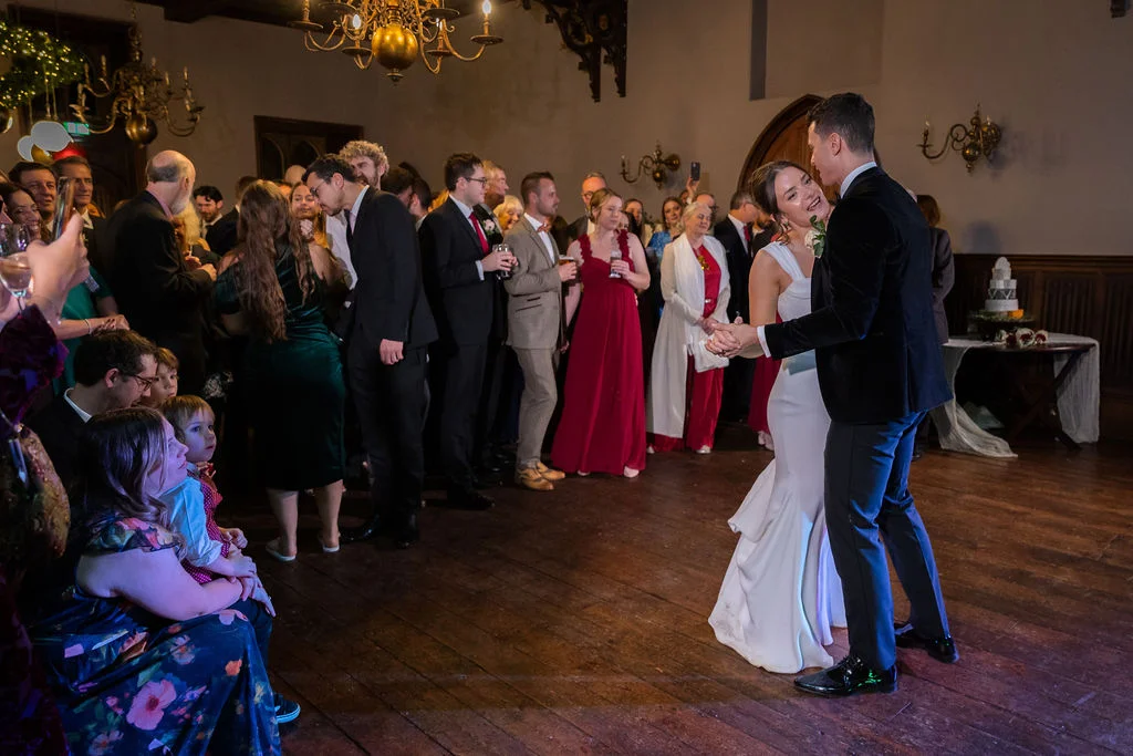 The bride and groom share their first dance on a wooden floor in a historic candlelit hall while guests watch closely.