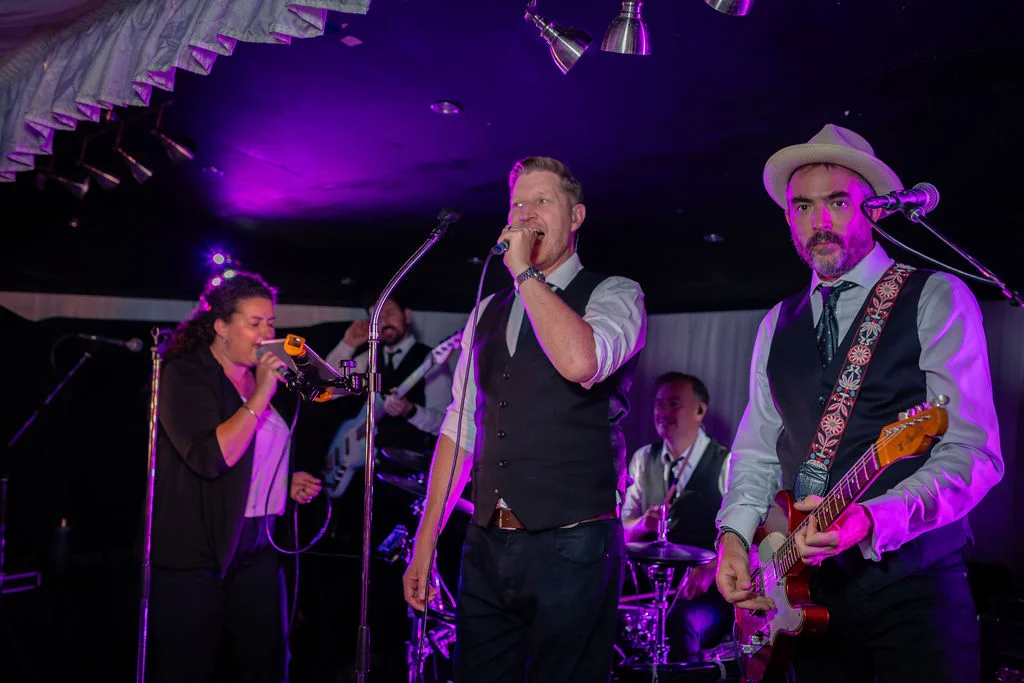 A lively wedding band performs on stage under dramatic purple lighting, featuring a male lead singer, a guitarist in a hat, and a female vocalist.
