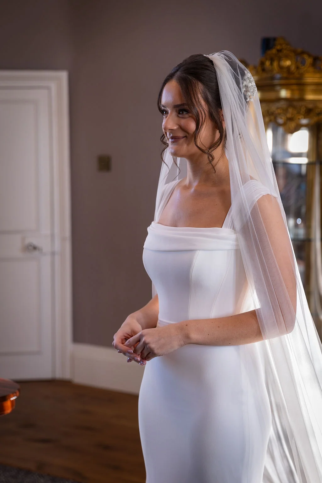 A smiling bride wearing a structured white crepe gown with a square neckline and a long veil stands indoors during bridal preparations.