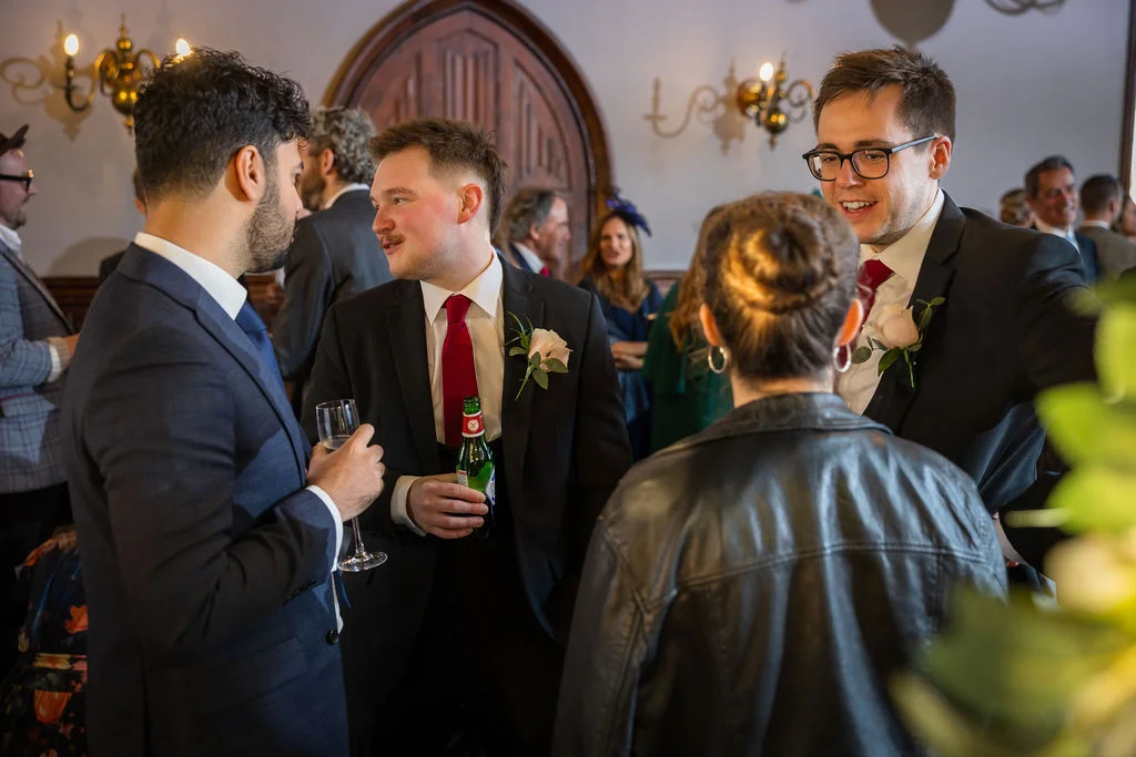 Groomsmen in dark suits and red ties mingle with guests during the wedding drinks reception in a hotel venue.