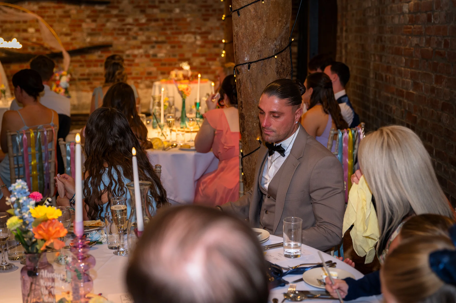 A well-dressed groom with a top knot and beige suit sits at a rustic barn wedding breakfast table surrounded by guests amidst exposed brick and candlelight.