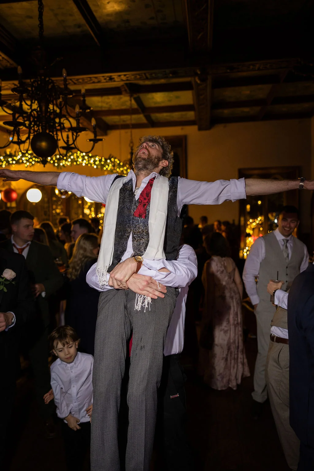 A groom is lifted high on the shoulders of wedding guests during a lively evening reception inside a dimly lit venue with chandeliers.