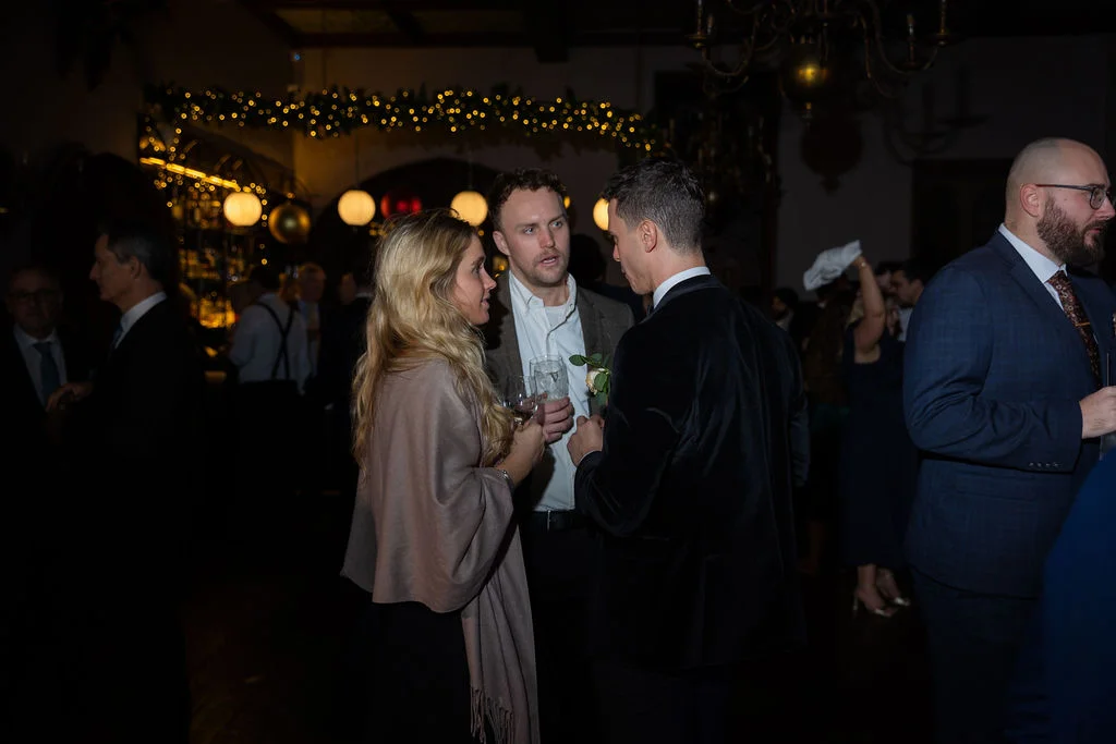 Wedding guests converse and mingle during an evening reception in a dimly lit ballroom adorned with warm fairy lights.
