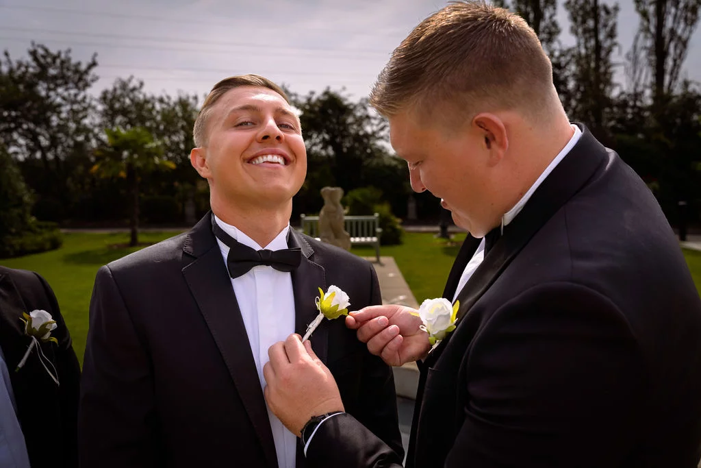 A groomsman wearing a black tuxedo smiles broadly as another person adjusts his white rose boutonniere in an outdoor garden setting.