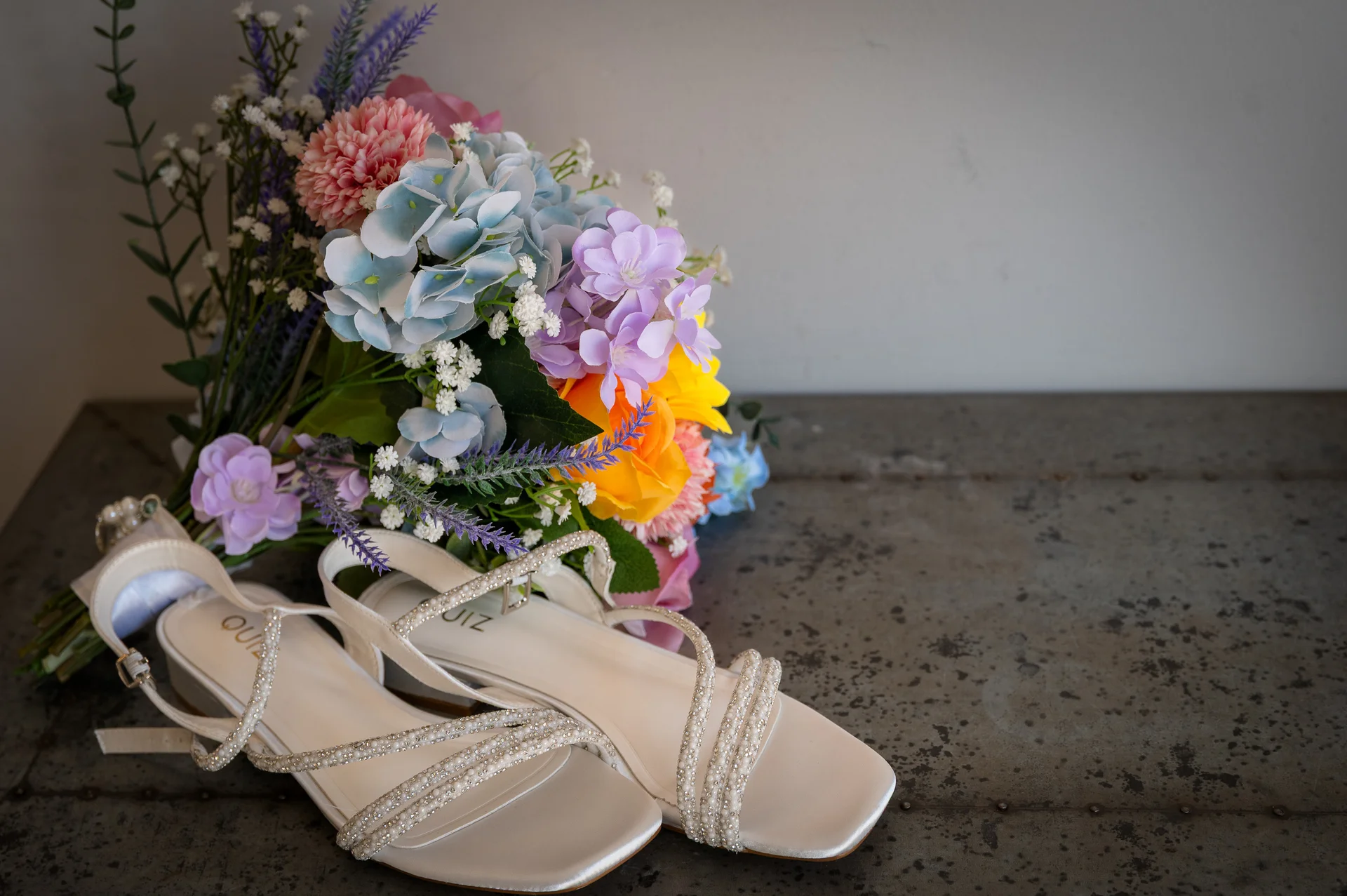 A flat lay detail of sparkling crystal-embellished strappy bridal heels resting next to a vibrant colourful wedding bouquet on a distressed metal surface.