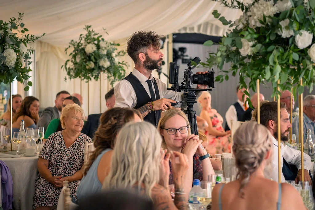 A videographer films wedding guests gathered inside a bright marquee decorated with tall floral arrangements during the wedding breakfast.