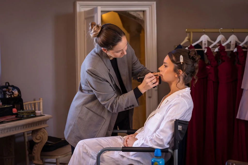 A professional makeup artist carefully applies lip liner to a bride sitting in a chair during morning preparations.