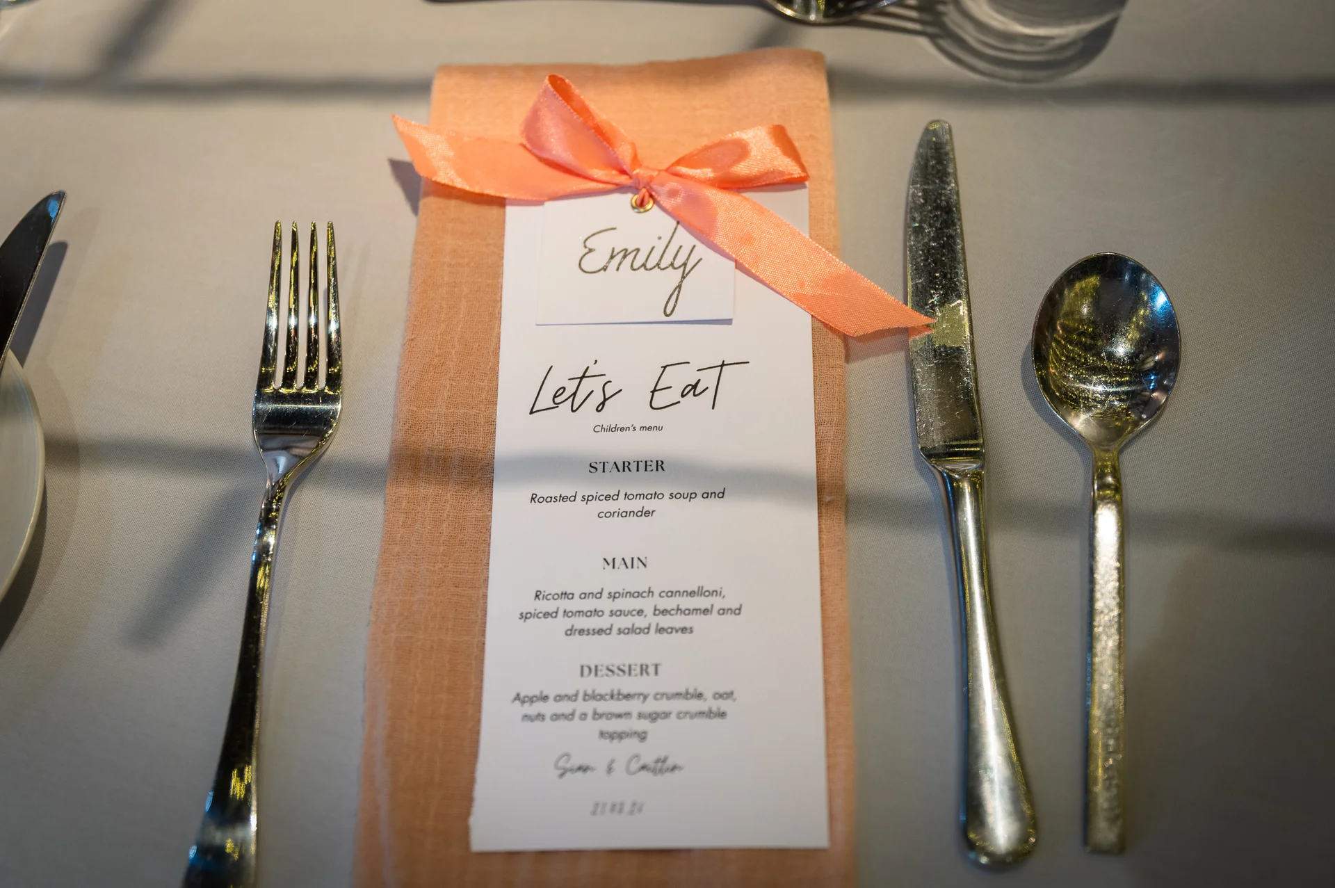 An elegantly styled wedding place setting features a peach napkin tied with a coordinating ribbon, a personalised menu card, and polished cutlery arranged neatly on a grey tablecloth.