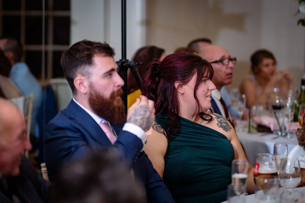 Wedding guests laugh and listen intently during speeches at a reception table set with drinks and glassware.