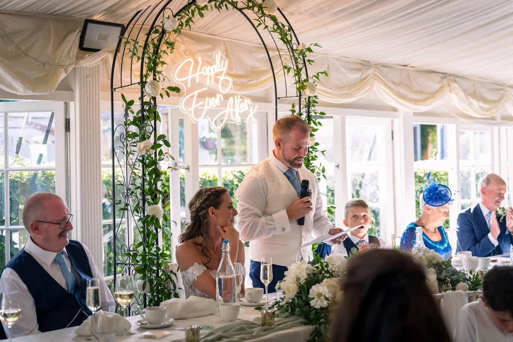 A groom delivers his speech while holding a microphone at the top table, standing next to his seated bride beneath a floral arch and a glowing neon sign.